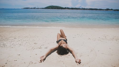 Sexy Tanned Girl with Beautiful Breasts, Lying on the Sand By the Ocean and Slowly Moves Her Foot