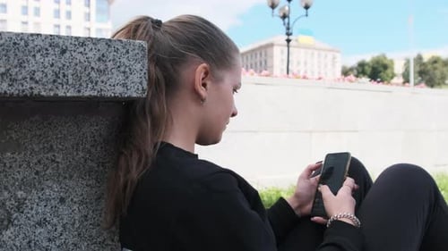 Young Woman Uses Smartphone While Sitting on the Parapet in the Street Park