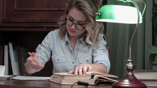 Beschäftigte Brillenstudentin, die mit einer grünen Lampe am Schreibtisch an ihrem Bericht arbeitet, Bücher durchgeht und