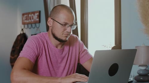 Smilling man freelancer in glasses and pink t-shirt working at a laptop in a coffee shop