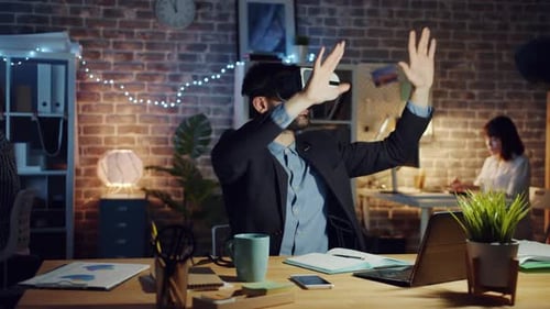 Man Using Virtual Reality Glasses Moving Arms Sitting at Desk in Dark Office