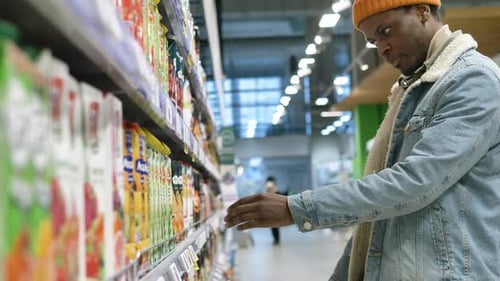 African American Guy in Hat and Jacket Chooses Juice in Shop