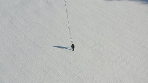 Birdseye aerial view of lonely man walking on snow capped field leaving trail on sunny winter day, h