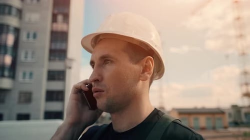 A Builder Wearing Helmet Is Talking By Phone While Inspecting Building Site.