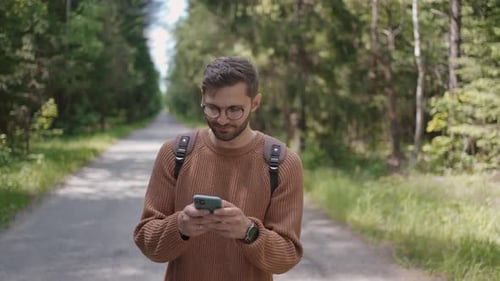 A Young Man with a Backpack Walks Along a Dirt Road in the Forest Writes Messages Communicates on