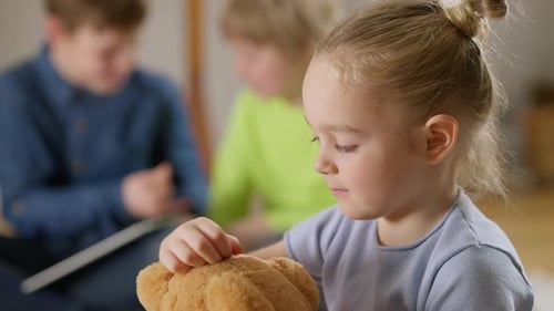 Closeup Side View of Little Pretty Girl Hugging Teddy Bear with Blurred Boys Talking at Background