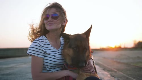 Happy Young Woman Holding and Hugging Her German Shepherd Dog Outdoor on the Sea Pier During Sunset