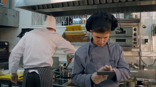 Young Cook Trainee Using Tablet While Chef Busy Cooking in the Kitchen