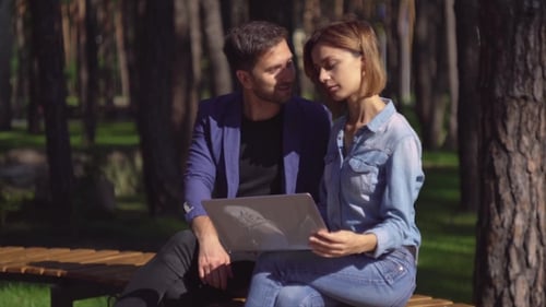 Young Couple Uses Laptop in Park on Wooden Bench