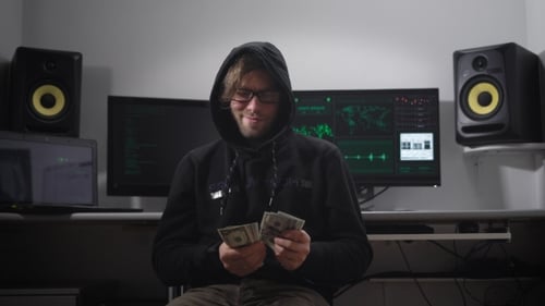 A Man in a Jacket with a Hood in the Computer Room. Hacker in Glasses and with Money in Hand Sits on
