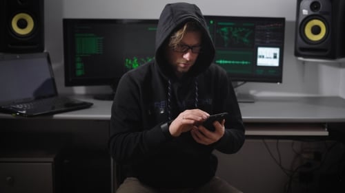 A Man in a Jacket with a Hood in the Computer Room. A Hacker in Sunglasses and with a Smartphone on
