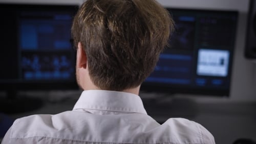 Experienced IT Specialist Works on a New Program on Two Computers. Young Man in Glasses and White