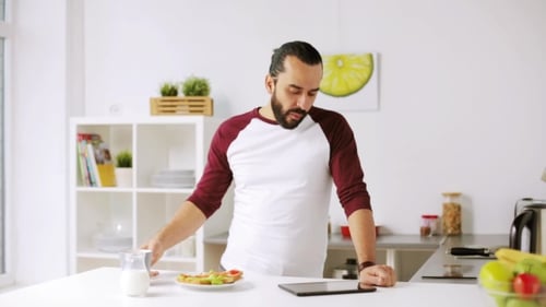 Man Drinking Coffee Looking at Tablet in Kitchen