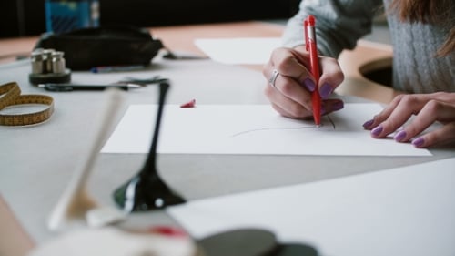 Woman Sitting at the Table in Office, Holding Pencil and Drawing Sketch of Shoes on Paper