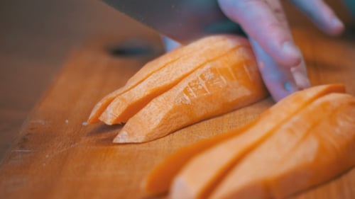 Woman Hands with a Knife Sliced Carrot with Knife on a Wooden Kitchen Board in a Home Kitchen