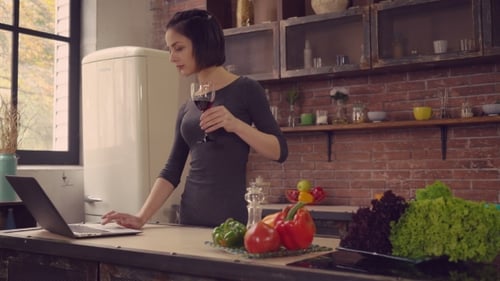 Woman Uses Laptop in Kitchen While Drinking Wine