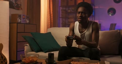 Um jovem estudante senta-se no sofá à noite na sala de estar segurando um console nas mãos brincando