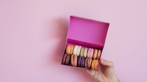 Hand Places Box of Macarons on Pink Background