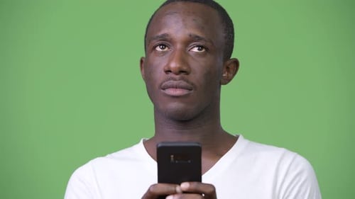 Young African Man Thinking While Using Phone Against Green Background