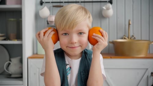 Smiling Boy Holds Oranges to Ears in Kitchen