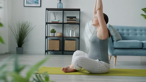 Side View of Attractive Expecting Mother Doing Yoga Stretching Body Sitting on Mat in Cozy Room