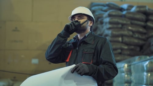 Engineer Studies Blueprint in Warehouse and Uses Radio