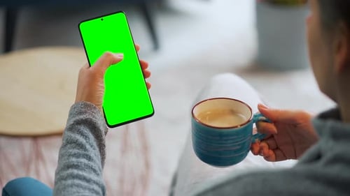 Woman at Home Using Smartphone with Green Mockup Screen in Vertical Mode and Drinking Coffee