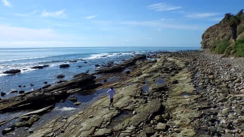 Tracking shot of a young man running on a rocky ocean beach shoreline