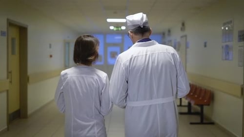 Back View of Male and Female Doctors Dressed in Lab Coats Walking Through the Corridor of the Ward