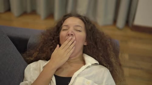 Schläfrige junge Frau gemischter Rasse gähnt auf grauem Sofa