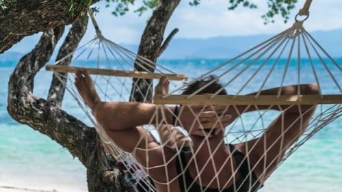 Man Swinging Relaxed in a Hammock on the Beach in Front of the Blue Ocean. Hiding Him From the Sun