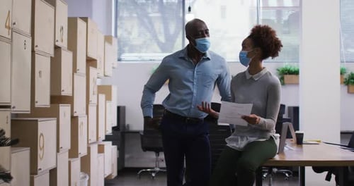 Diverse male and female business colleagues in face masks talking, woman holding document in office
