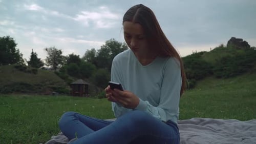 A Young Girl Sitting on Blankets on the Lawn in the Park and Gaining a Message on the Phone