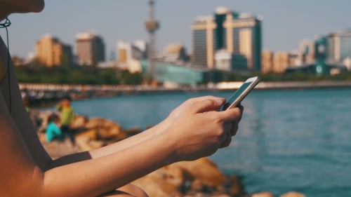 Young Woman Using Mobile Phone Sitting on Embankment Near the Sea and Skyscrapers of Megapolis