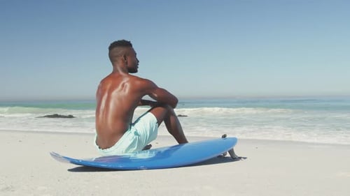African American man sitting at beach with surfboard