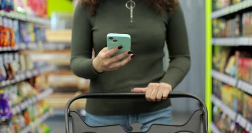 Close Up of Unrecognizable Woman Using Smartphone While Carrying Shopping Cart Walking Through