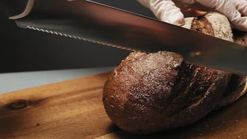 Gloved Hand Slices Brown Bread on Wooden Board