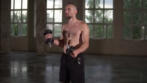 Muscular Man Curls Dumbbells in Empty Warehouse