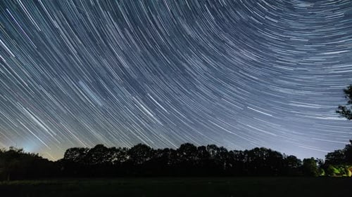moving night sky with star trails over rural landscape with field in front of trees