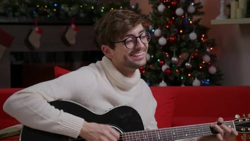 Man close up playing Christmas song on guitar near decorated Christmas tree.