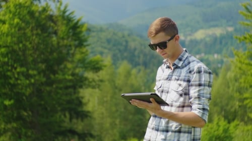 Un joven usa una tableta, se para en un lugar pintoresco sobre un fondo de hojas verdes
