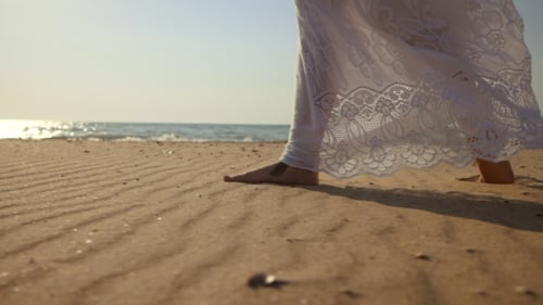 Legs of Caucasian Girl Wearing White Long Dress and Silver Bracelets Walking Barefoot Sand on Sea