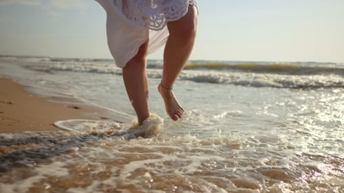 Legs of Caucasian Girl Wearing White Long Dress and Silver Bracelets Walking Barefoot Sand on Sea