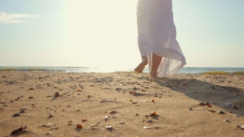 Legs of Caucasian Girl Wearing White Long Dress and Silver Bracelets Walking Barefoot Sand on Sea