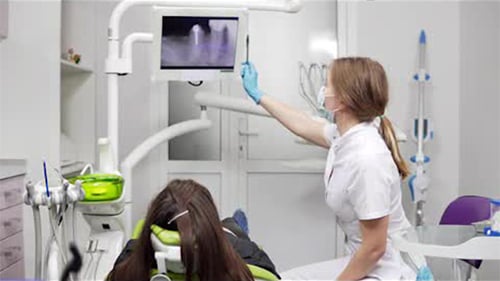 Junge Zahnärztin zeigt am Stuhl Röntgenzähne auf einem Tablet. Zahnärztin in Maske und Labor