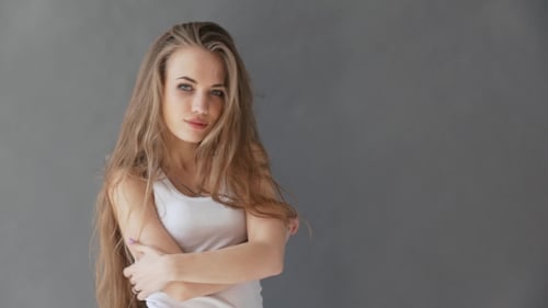 Belle jeune femme aux cheveux blonds sains. Dans un débardeur blanc sur fond gris, posant sur