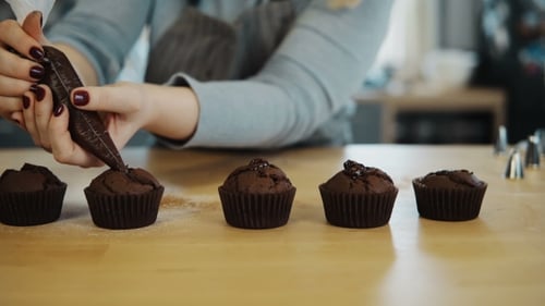 Blick auf weibliche Hände legt die Creme aus dem Spritzbeutel auf Schokoladen-Cupcakes Junge Frau kocht