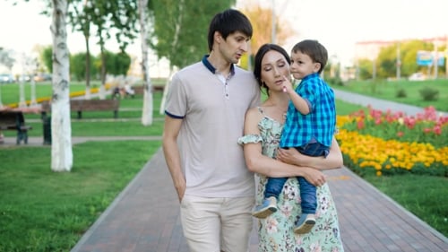 Happy Young Family Is Strolling in a Park in Summertime, Mother Is Holding Her Little Son