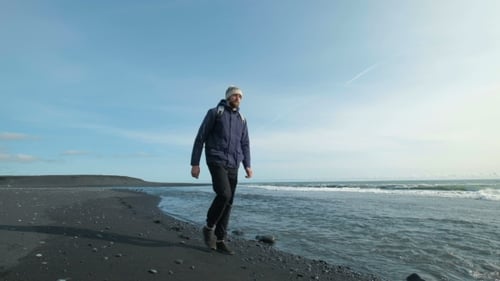Young Man Walking on a Black Sandy Beach on a Mountain. During Vacation the Man Is in Warm Clothes