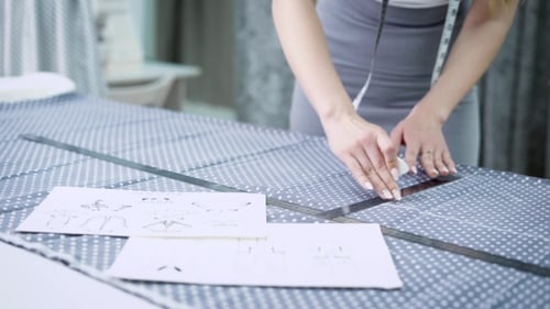 of Young Female Designer Standing at the Table in Tailoring Studio Making Lines on Cloth with Chalk